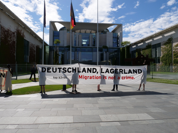 Eine Gruppe von Menschen, die ein Transparent mit der Aufschrift "Deutschland, Lagerland Migration ist kein Verbrechen" vor einem umzäunten Gebäude mit Bäumen und Flaggen hölt.