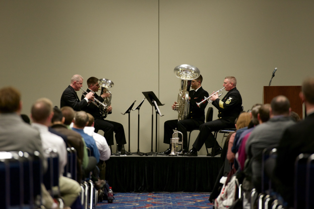 Eine Gruppe von Männern, die auf einer Bühne Blasinstrumente spielen, mit Notenständern, einem Podium und einem Mikrofon vor einem Publikum, das auf Stühlen sitzt.