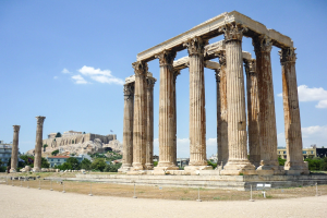 Alter Tempel von Olympie Zeus in Athen, Griechenland, mit hohen Säulen, umgeben von Gebäuden, Bäumen und Felsen, mit einer Festung in der Ferne im Hintergrund unter einem bewölkten Himmel.
