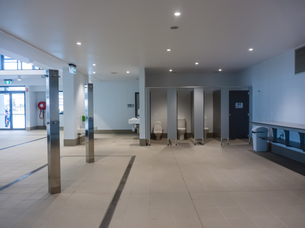 Große Öffentliche Toilette mit mehreren Toiletten, tragenden Säulen, einem Mülleimer, einer Tür, einem Schild, einem Feuerlöscher und Deckenleuchten.