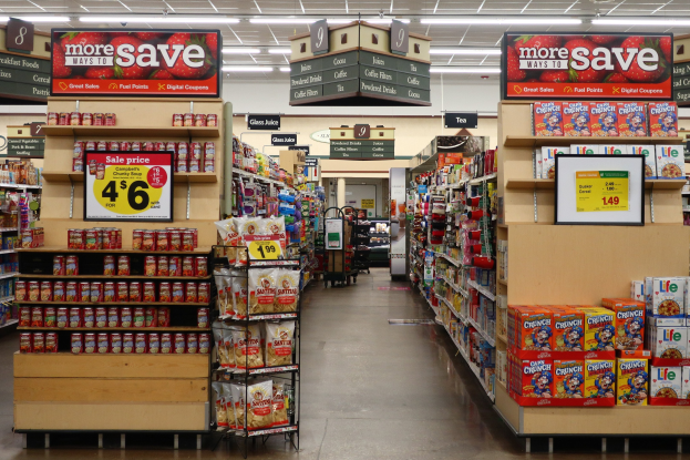 Ein Kiosk-Innenraum Mit Regalen Und Boden, Der Mit Verschiedenen Lebensmitteln Bestückt Ist, Beleuchtung Von Oben Und Ein Numeriertes Schild, Mit Einem Menschen Im Hintergrund.