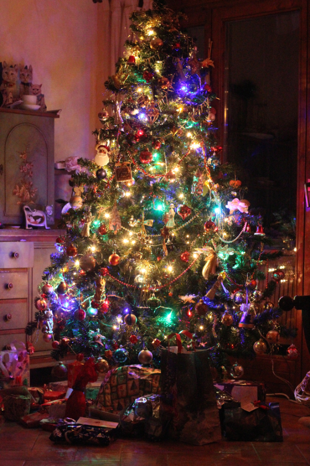 Ein Weihnachtsbaum mit Lichtern und Deko, Geschenke auf dem Boden, ein Schrank mit Gegenst√§nden links und ein Fenster an der rechten Wand.