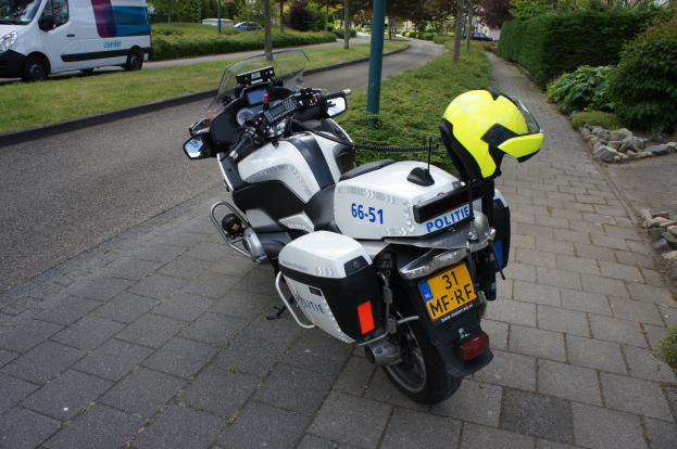 Ein Polizeimotorrad am Straßenrand geparkt mit einem Helm auf dem Sitz, umgeben von Bäumen, Gebäuden und anderen Fahrzeugen.