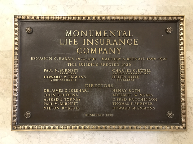 Plakette an einer Gebäudewand mit der Aufschrift "Monumentale Lebensversicherungsgesellschaft" in fetter Schrift, die in einem dekorativen Rahmen auf dunklem Material gesetzt ist.