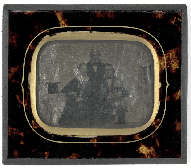 Schwarze-und-weiß-Foto eines Mannes in Anzug und Krawatte und einer Frau in unbekannter Kleidung, die vor einem Fernseher stehen, beide mit ernsten Gesichtern.