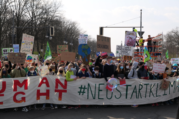 Eine große Gruppe von Menschen marschiert auf einer Straße, hält eine 'Menschliche Rechte' Fahne und verschiedene Plakate, während einige Masken tragen, vor einem Hintergrund aus Bäumen, Laternenpfählen und einem klaren blauen Himmel.