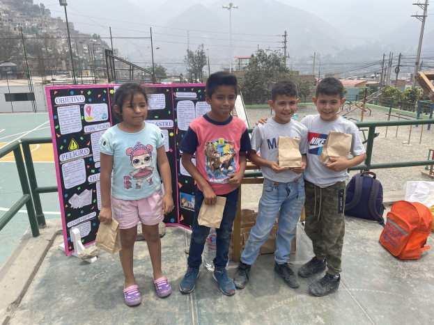 Gruppe von Kindern mit Papiertüten auf einem Basketballfeld mit Tüten und Gegenständen auf dem Boden, eine Tafel mit Text links daneben und Zaun, Pfosten, Straßenlaternen, Stromleitungen, Bäume, Gebäude, Hügel und Himmel im Hintergrund.
