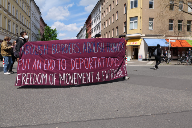 Eine Gruppe von Menschen marschiert mit einem Banner, auf dem "Abolish Borders, Abolish Frontiers, Put an End to Deportations, Freedom of Movement 4 Everyone" steht, die Straße entlang vor Gebäuden, Bäumen und Fahrrädern unter einem bewölkten Himmel.