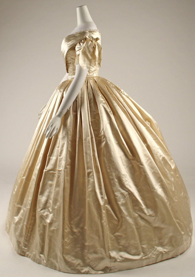 Eine Schaufensterpuppe in einem Museum präsentiert ein goldenes Ballkleid mit hohem Ausschnitt, langen Ärmeln und einem voluminösen Rock mit aufwendigen Details und einer eng anliegenden Taille.
