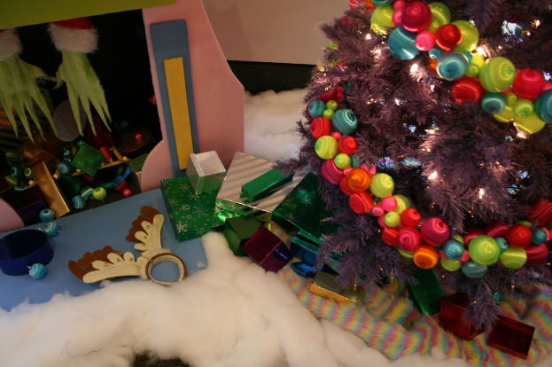 Eine Indoor-Szene mit einer Wand im Hintergrund, einem Baumwoll- und Wollstoff am Boden, einem geschmückten Weihnachtsbaum auf der rechten Seite, Geschenkboxen und Dekorationsgegenständen in der Mitte und einer Hütte mit Dekorationen und Baumwolle oben links.