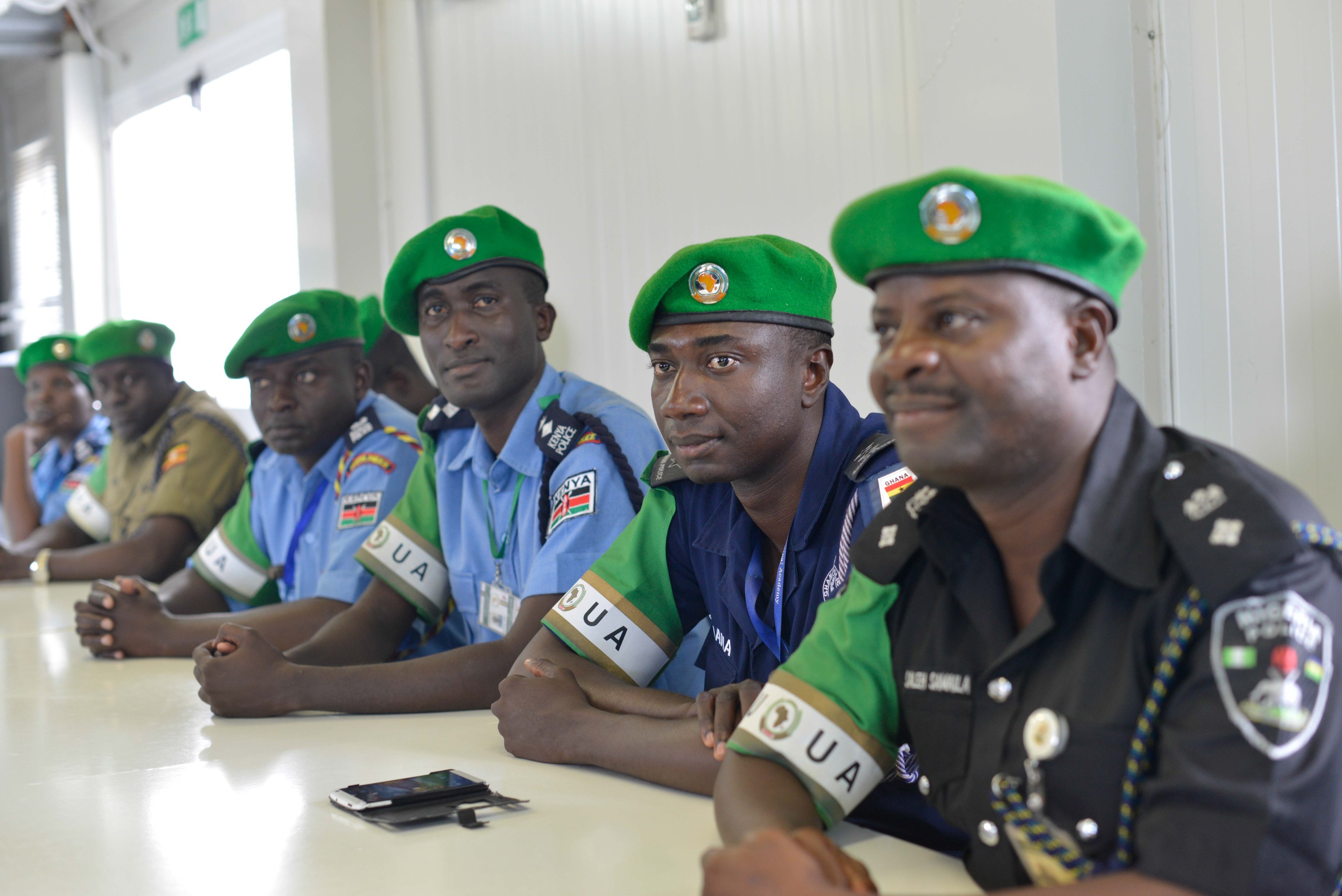 Eine Gruppe von Polizeibeamten sitzt an einem Tisch während einer Schulung, mit Mobiltelefonen und anderen Gegenständen auf dem Tisch, in einem Raum mit Fenstern und Deckenbeleuchtung.