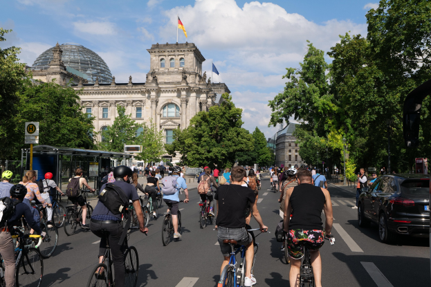 Gruppe von Menschen, die auf Fahrrädern eine von Bäumen gesäumte Straße in Berlin entlangfahren, mit Gebäuden und einer Bushaltestelle, unter einem bewölkten Himmel und einer Flagge an einem Gebäude.