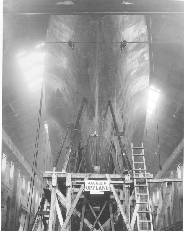 Großes Schwarz-Weiß-Foto eines großen Bootes im Bau in einer Werft, mit einer Leiter, Holzpfählen, einem Schild, Metallrahmen, Deckenleuchten und Fenstern.