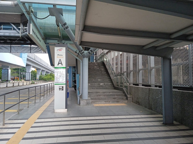 Ein Bahnsteig mit Treppe, eine Straße mit Geländer auf der linken Seite und ein Hintergrund mit Bäumen, Pfählen, Gebäuden und einer Brücke, mit Rohren und einer Tafel mit Text oben auf dem Bahnsteig.