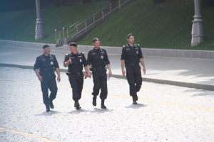 Drei Polizisten in Uniform und Mützen gehen auf einer Straße mit Pfählen, Geländern, Treppen und Gras im Hintergrund.