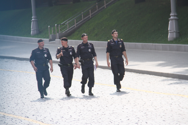 Drei Polizisten in Uniform und Mützen gehen auf einer Straße mit Pfählen, Geländern, Treppen und Gras im Hintergrund.