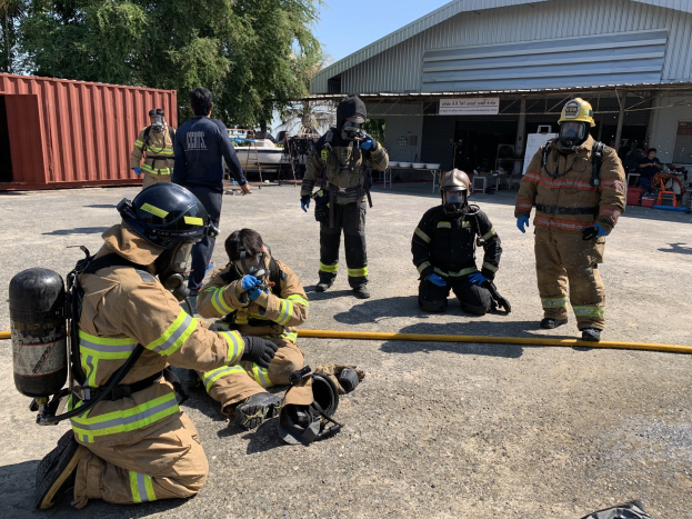Feuerwehrleute mit Helmen und Gasmasken sitzen während einer Übung vor einem Gebäude auf dem Boden, mit einem Rohr im Vordergrund, einem Container links, Bäumen und einem Schuppen im Hintergrund und dem Himmel darüber.
