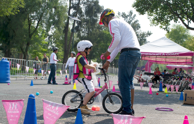 Ein Mann hilft einem jungen Mädchen beim Fahrradfahren in einem Park, wobei ein pinkes und weißes Kleid und ein weißer Helm am Mädchen zu sehen sind.