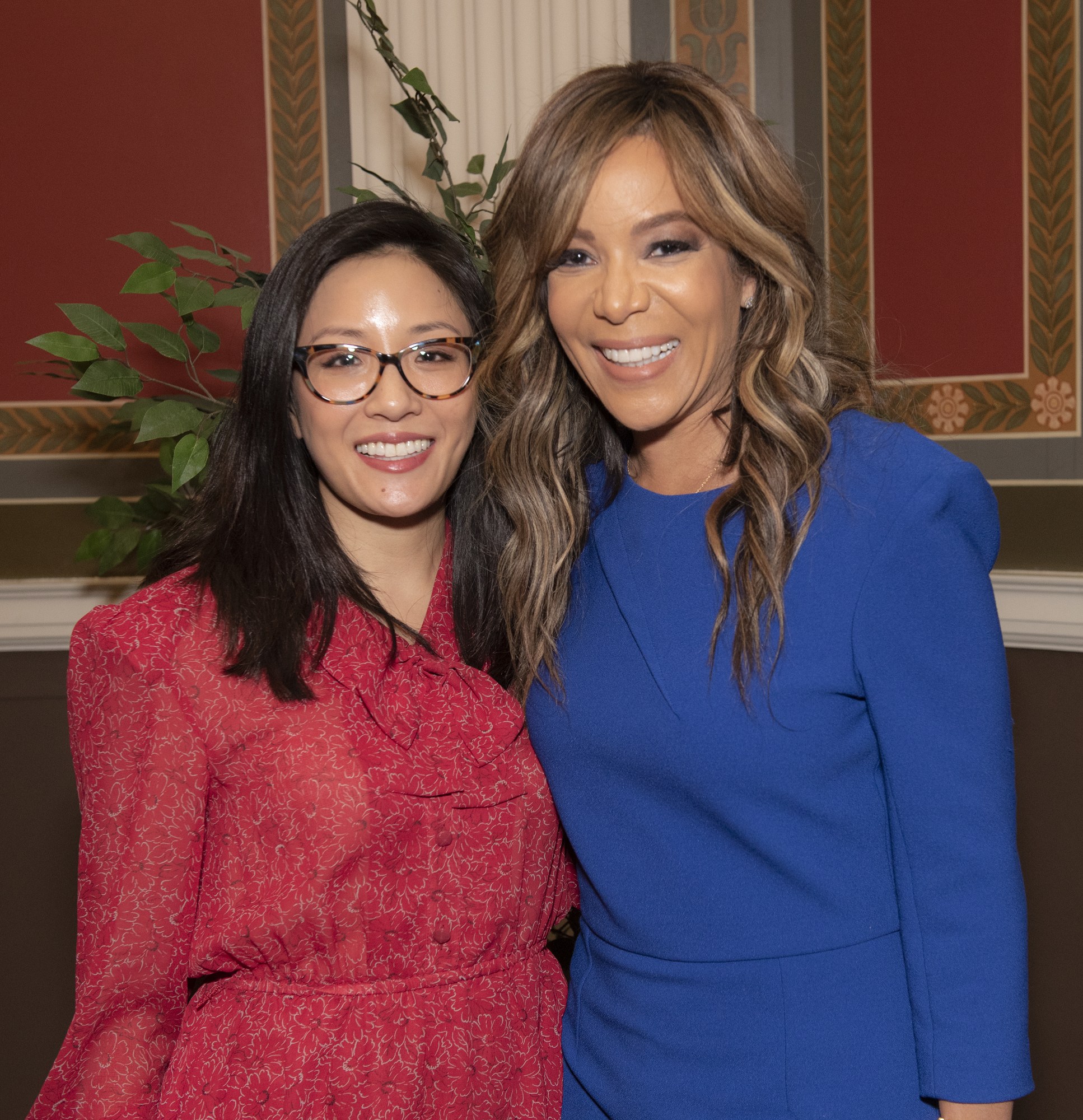 Zwei Frauen in Abendgarderobe stehen strahlend nebeneinander bei einem Event, eine in einem blauen Kleid und die andere in Rot mit Brille, mit einer Pflanze und einem Vorhang im Hintergrund.
