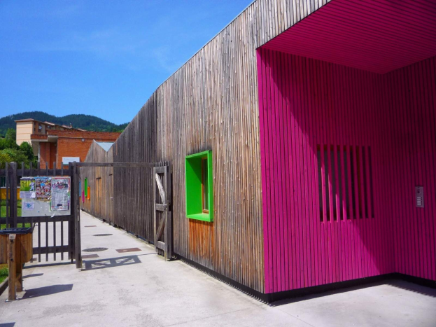 Pinkes Gebäude mit grüner Tür und Fenstern, hölzerner Zaun, Anschlagtafel mit Papieren, Mülltonne, Gras, Bäume, entfernte Gebäude, Hügel und bewölkter Himmel, Teil eines neu transformierten Schulspielplatzes in Spanien.