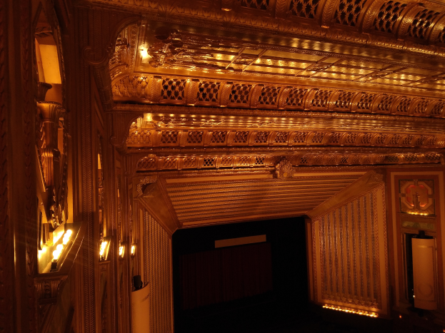 Ein prunkvolles Theaterinnere mit dunklem Hintergrund, beleuchtet von Lichtern auf beiden Seiten, mit einer kunstvoll gestalteten Decke.