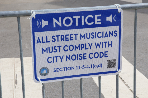Hinweistafel an einem Zaun mit einem Schild, das Straßenmusikern die Einhaltung der städtischen Lärmvorschriften vorschreibt, mit einer Straße im Hintergrund.