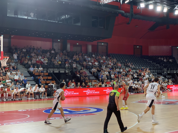 Eine Gruppe von Menschen, die Basketball in einer Halle spielen, mit einem Basketballkorb auf der linken Seite und Zuschauern im Hintergrund.