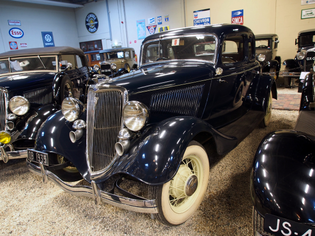 Eine Gruppe von Oldtimern in einer Garage geparkt, mit einer Wand im Hintergrund, die mit Brettern und Leuchten an der Oberseite verziert ist, die den Eindruck eines klassischen Autotreffens erweckt.