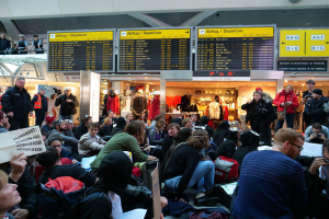 Eine große Gruppe von Menschen sitzt und steht in einem Flughafen während einer Protestaktion, wobei einige Taschen und Papiere halten, während Schilder mit Text, Puppen in Kleidern und Deckenleuchten im Hintergrund zu sehen sind.