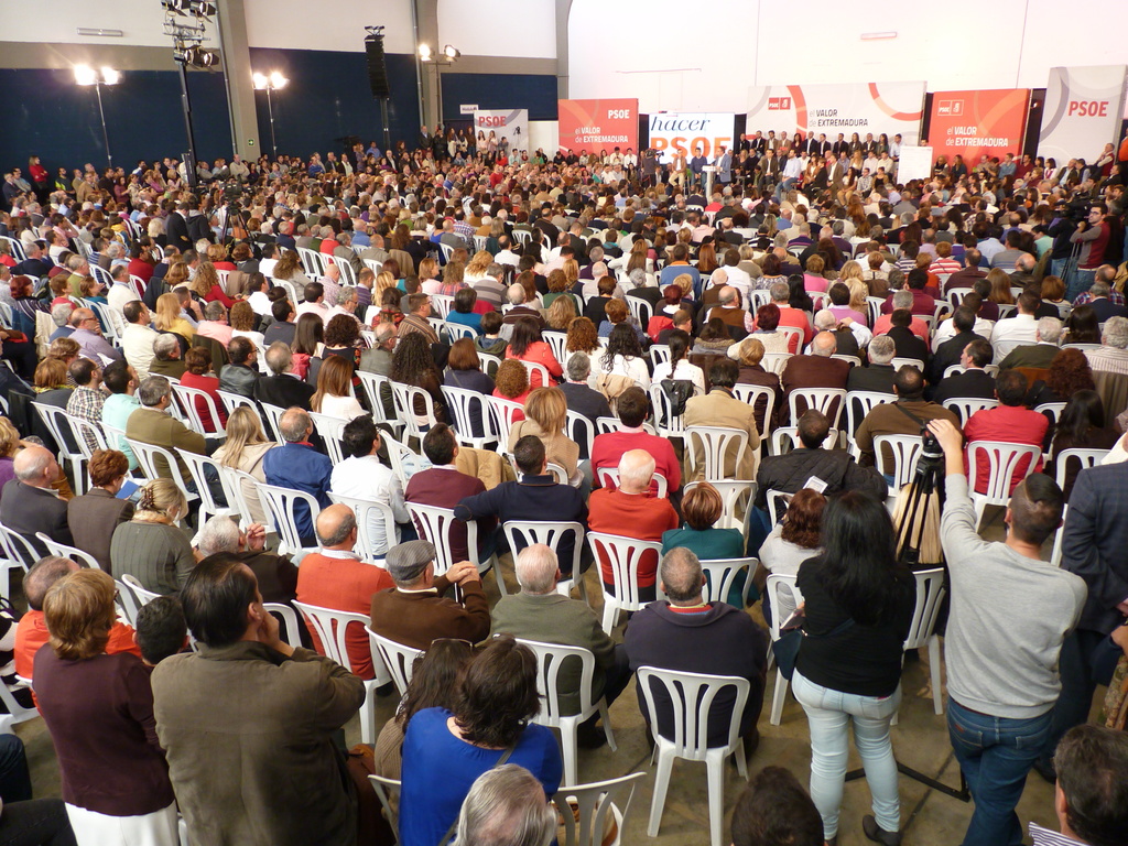 Ein volles Stadion mit vielen sitzenden und stehenden Menschen, eine Bühne mit zwei Rednern an einem Podium und zahlreichen Fahnen und Lichtern.