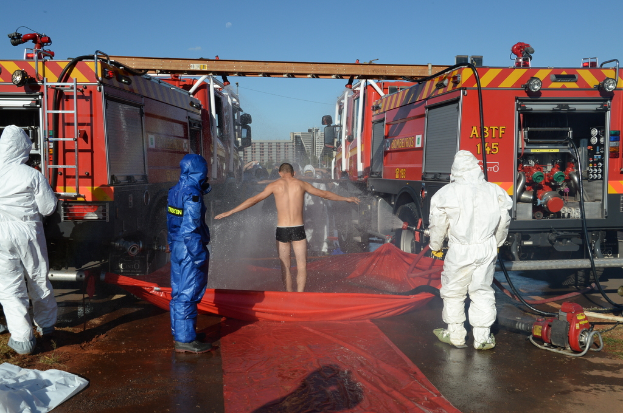 Drei Personen in Warnwesten stehen im Vordergrund, eine Person steht auf einer Abdeckung über Wasser vor ihnen. Einige andere sind in der Nähe, und Feuerwehrautos mit ausgefahrenen Metallstangenleitern sind daneben positioniert. Gebäude sind im Hintergrund zu sehen, und es gibt verschiedene Gegenstände auf der Oberfläche.