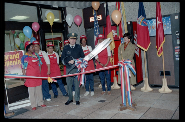 Eine Gruppe von Menschen versammelt sich vor einem Laden, waehrend ein Mann in der Mitte eine Schnur mit einer Schere schneidet.