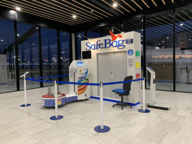 Sicherheitskontrolle am Flughafen mit einem Stuhl, Absperrpollern, Maschinen, einem Bildschirm, einer bedruckten Tafel, Glaswänden, Deckenleuchten und sichtbarem Boden.