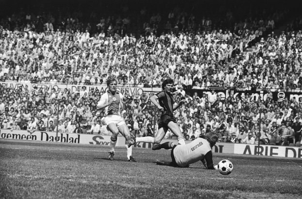Schwarzes Foto von Männer, die auf einem Feld Füßball spielen, mit Zuschauern im Hintergrund und einer 'Bundesliga 1978-79 Ajax 1-1 Hannover 96'-Fahne.