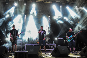 Drei Männer auf der Bühne, die Gitarren spielen und in Mikrofone singen, umgeben von Musikausrüstung und einem Banner mit Text und Lichtern im Hintergrund beim Belgrade Music Festival in Serbien im Jahr 2016.