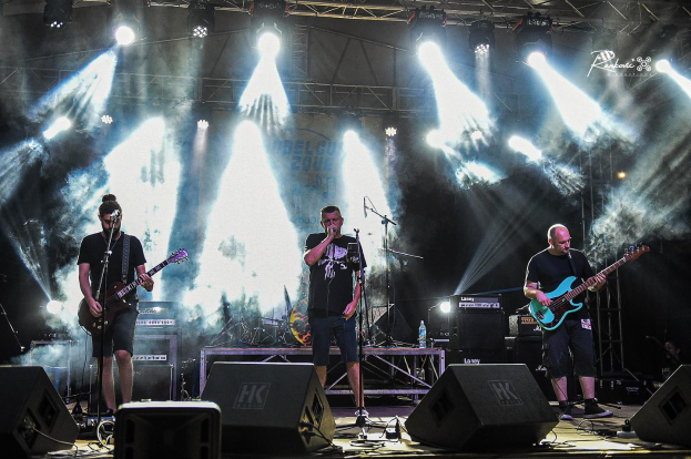 Drei Männer auf der Bühne, die Gitarren spielen und in Mikrofone singen, umgeben von Musikausrüstung und einem Banner mit Text und Lichtern im Hintergrund beim Belgrade Music Festival in Serbien im Jahr 2016.
