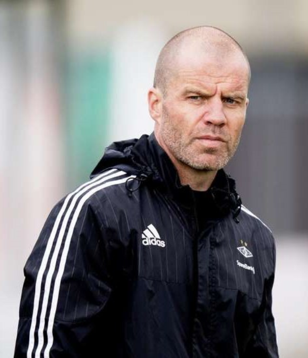 Kahlköpfiger Trainer in marineblauer Jacke mit weißen Streifen und weißem Kragen auf einem Fußballfeld mit gefalteten Händen und ernster Miene.