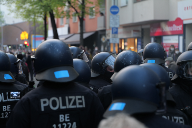 Eine Gruppe von Polizisten in schwarzen Uniformen und Helmen steht vor einer unscharfen Menge und einer Stadtlandschaft mit Geb├Ąuden, B├Ąumen, Masten, Verkehrszeichen und Schildern mit Text.