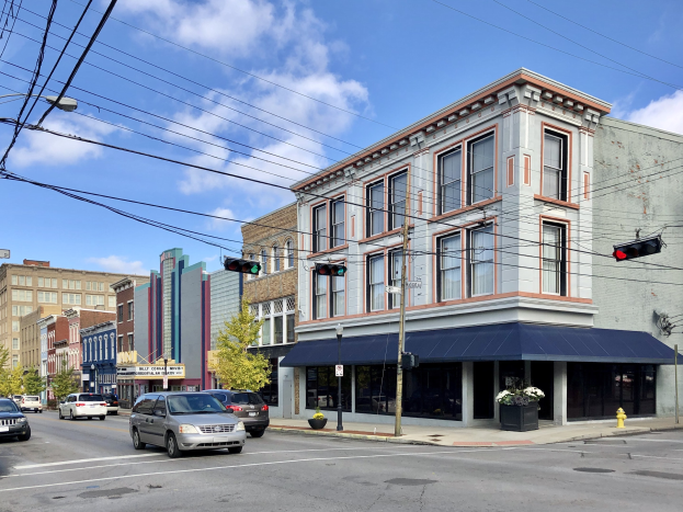 Eine Stra├če in einer Stadt mit parkenden Autos, Strommasten, Ampeln, Stra├čenlaternen, B├Ąumen und Geb├Ąuden mit Fenstern, unter einem bew├Âlktem Himmel, mit Text, der unten eine gewerbliche Immobilie zum Verkauf f├╝r 1.250.000 USD anzeigt.