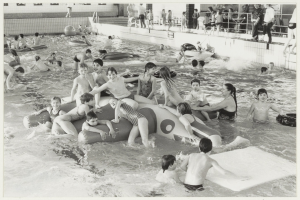 Schwarzes und weißes Bild von Kindern, die in einem Schwimmbad spielen, einige auf Schwimmhilfen, andere stehen, mit einer Wand und einem Geländer im Hintergrund.