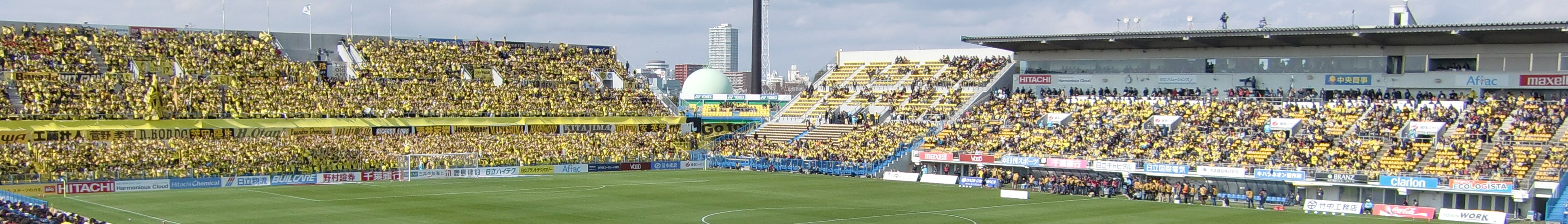 Großes Stadion voller Zuschauer bei einem Fußballspiel auf saftigem Grün, umgeben von Werbetafeln mit Text, mit Gebäuden, Pfählen und einem klaren blauen Himmel im Hintergrund.