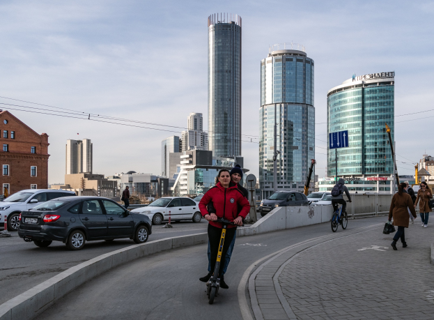 Ein Mann in einer roten Jacke und schwarzen Hosen fährt auf einem Elektroroller die Straße mit hohen Gebäuden hinunter, umgeben von Passanten und Radfahrern unter einem teilweise bewölktem Himmel.