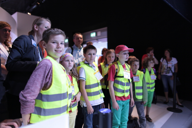 Eine Gruppe von Kindern in Sicherheitswesten und Namensschildern steht auf einem Boden, mit einem Jungen auf der linken Seite, der lächelt, einem Pfahl auf der rechten Seite und ein paar Leuten im Hintergrund.