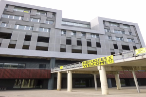 Großes Gebäude mit einem "Willkommen bei den Flüchtlingen"-Schild, umgeben von Säulen und einem Geländer, unter einem sichtbaren Himmel.