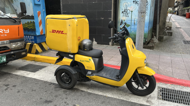 Ein gelbes Elektro-Scooter mit Helm steht auf der Straße neben einem Lastwagen, mit Gebäuden und Passanten im Hintergrund.