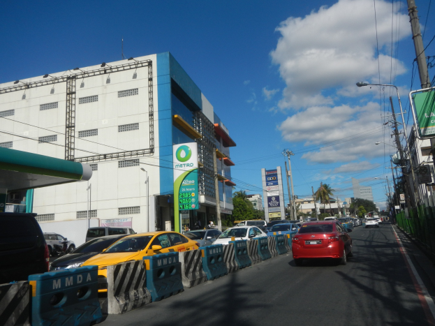 Eine belebte Stadtstraße mit hohen Gebäuden, mehreren Fahrzeugen, Absperrungen, Schildern, Laternenmasten, Stromleitungen, Bäumen und einem bewölkten Himmel.