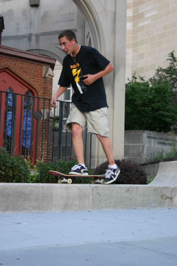 Ein Mann, der auf einer vertikalen Wand Skateboard fährt, mit einer Plattform unten, einem Haus, einem Grill, Pflanzen, Bäumen, einer Backsteinwand und einer Säule im Hintergrund.