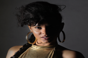 Eine Frau in einem goldenen Kleid und großen Ohrringen steht selbstbewusst vor einer Wand, ihr glatter Bob-Haarschnitt und das natürliche Make-up unterstreichen ihren entschlossenen Gesichtsausdruck, während sie direkt in die Kamera blickt.