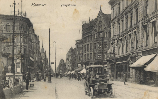 Ein altes Schwarz-Weiß-Foto einer Straßenkreuzung in Hannover, Deutschland, mit Fahrzeugen auf der Straße, Fußgängern auf den Gehwegen, Gebäuden mit Fenstern auf beiden Seiten, Pfählen, einer Uhr und einem sichtbaren Himmel.