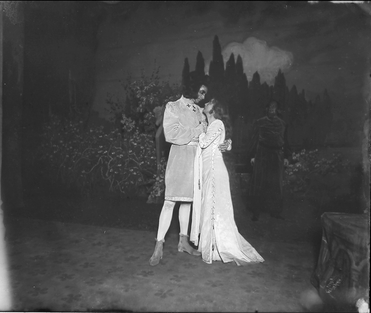 Ein Schwarz-Weiß-Foto eines Mannes und einer Frau, die sich auf einer Bühne umarmen und küssen, mit einem Publikum, Pflanzen und Bäumen im Hintergrund.